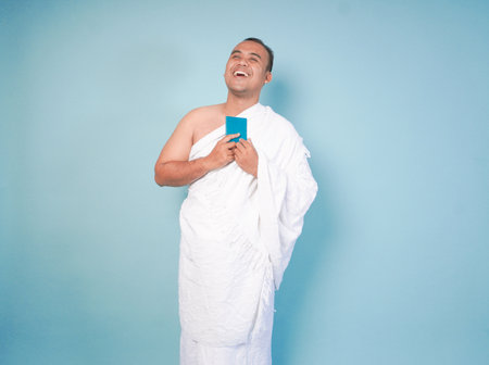 young Asian Muslim man wearing ihram posing while holding passpor isolated blue background. Hajj...