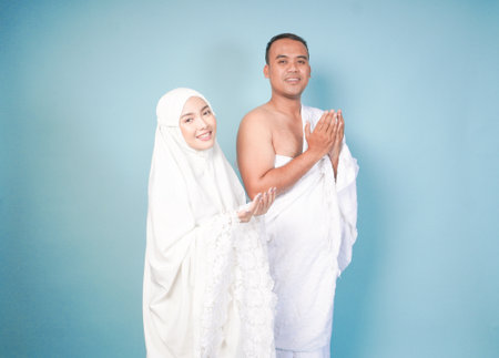 young Asian Muslim couple wearing ihram and prayer gown doing praying gesture over isolated by blue...