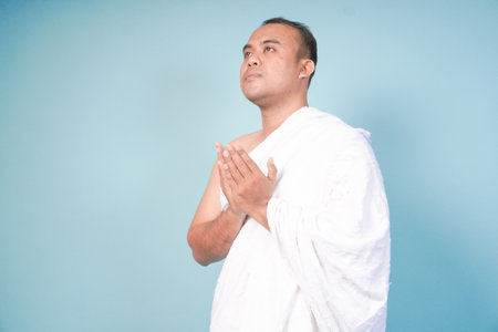 young Asian man Muslim wearing ihram and prayer gown doing praying gesture over isolated by blue...