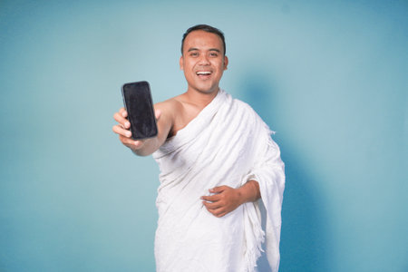 young Excited Asian Muslim man wearing ihram pointing to copy space on his smartphone, isolated blue...