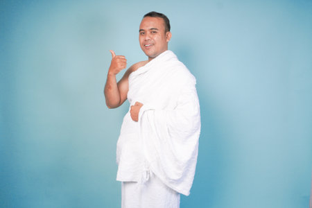Excited Asian Muslim man wearing ihram, gesture thumbs up pose, isolated blue background. Hajj...