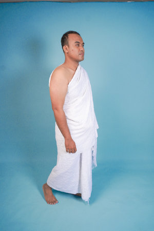 Handsome young Asian Muslim man wearing ihram walking over isolated by blue background. Hajj...