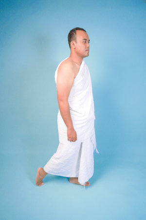 Handsome young Asian Muslim man wearing ihram walking over isolated by blue background. Hajj...