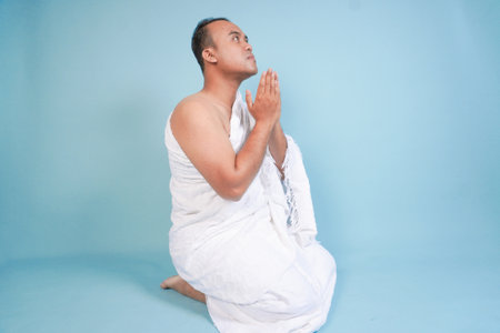 Young serious Asian Muslim man wearing ihram sitting and praying holding hands up with hand gesture isolated on blue background. Hajj concept. islam muslim conceptの写真素材