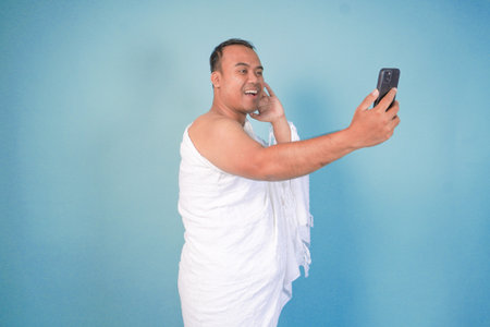 young Asian Muslim man wearing ihram doing video call pose while holding smartphone, isolated blue...