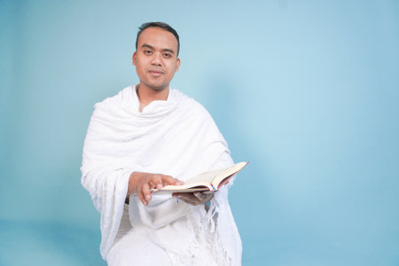 Serious young Asian Muslim man wearing ihram sitting praying worship reading al quran isolated on...