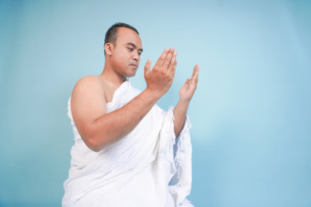 Young serious Asian Muslim man wearing ihram sitting and praying holding hands up with hand gesture...