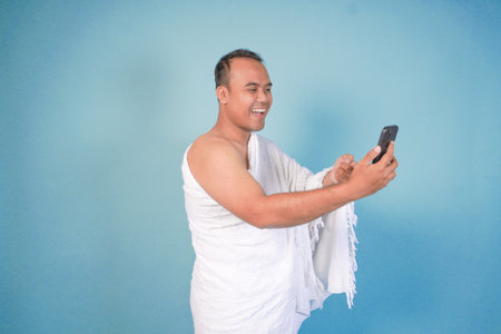 young Asian Muslim man wearing ihram doing video call pose while holding smartphone, isolated blue...