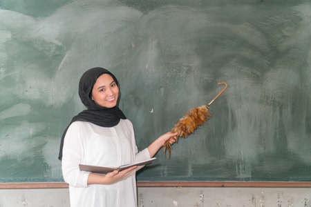 Malay School Teacher wearing hijab holding book and feather duster