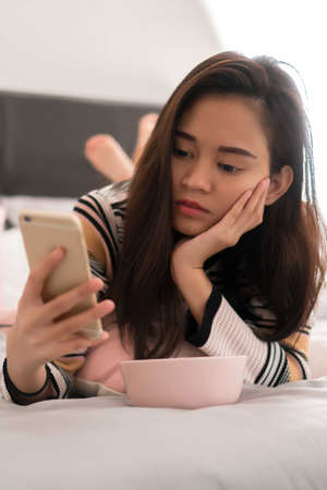 Beautiful young Asian girl on the bed holding mobile phoneの写真素材