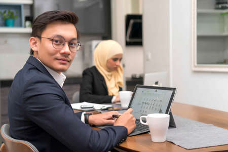 Asian malay man working at home with laptop with colleague at backgroundの写真素材