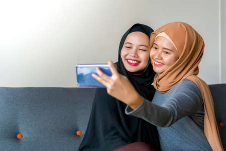 Two Malay friends wearing hijab at the sofa using smart phone taking selfieの写真素材