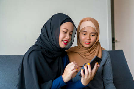 Two Malay friends wearing hijab at the sofa using smart phone