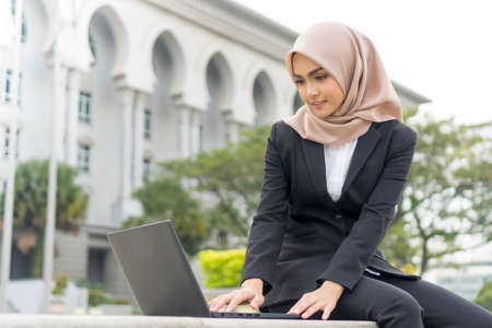Cute Malay Woman wearing hijab outdoor sitting and using laptopの写真素材