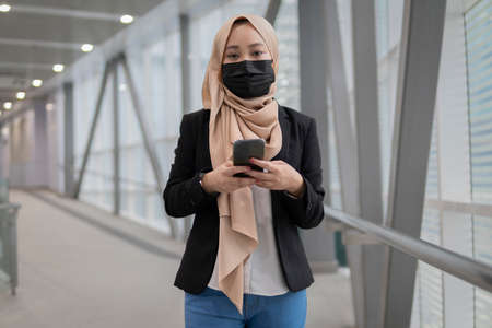 New Normal Concept: Young malay lady wearing face mask holding smartphone