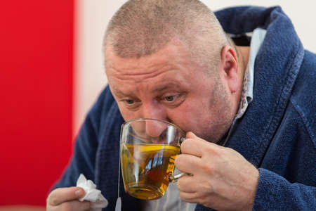 Ill man blowing his nose with paper napkin and drinking tea at homeの写真素材