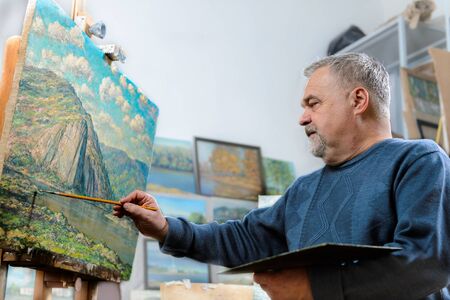 Elderly artist paints a picture oil paints in his studio
