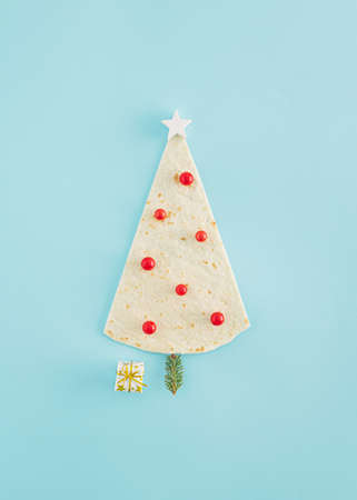 Creative Christmas tree idea made of a tortilla or pizza dough, a white wooden star, pine twig, a gift and red baubles on a blue background. Minimal flat lay, New Year concept. Christmas food inspiration.の写真素材