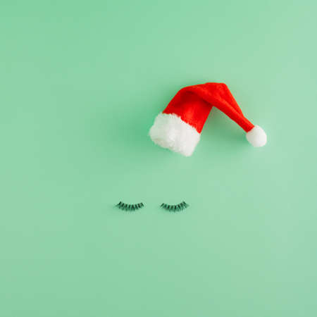 Creative Christmas arrangement made of eyelashes and red Christmas cap on a pastel green background. Minimal New Year flat lay concept. Christmas dream and Santa face inspiration.の写真素材