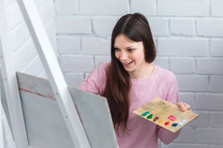 Beautiful young woman painting a picture on an easel. Hold in your hands a brush and palette of paints.の写真素材