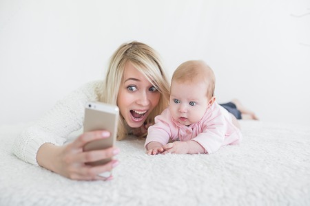 Funny baby girl make selfie on mobile phone and lying near her mother.の写真素材
