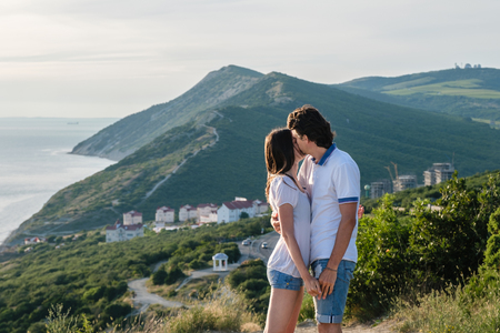 Man kisses a woman on the cheek and hugs, standing on a mountain with seascape. Side view.の写真素材
