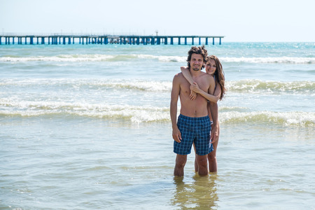 Brunette woman in bikini hugs a beautiful long-haired man from behind, standing in the sea.の写真素材