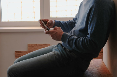 Man with phone. Unrecognizable young man playing games in mobile phone sitting on balcony near the windowの写真素材