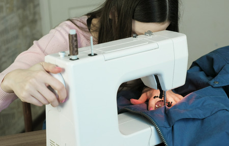 Young woman sews blue jacket on white sewing mashine.
