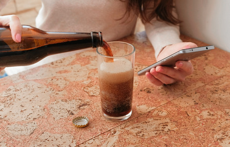 Young beautiful girl reads something on the phone, pours beer into a glass. Closeup hands.の写真素材