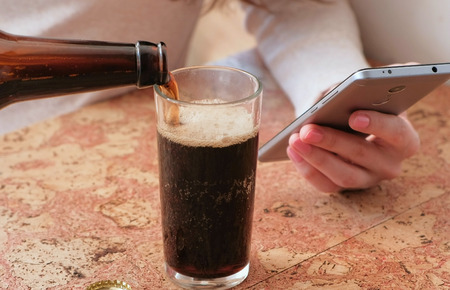 Young beautiful girl reads something on the phone, pours beer into a glass. Closeup hands.