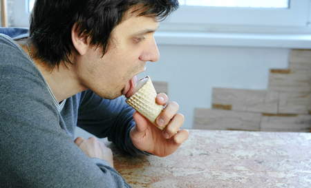 Young man eats ice cream at home sitting by the window. Side view.