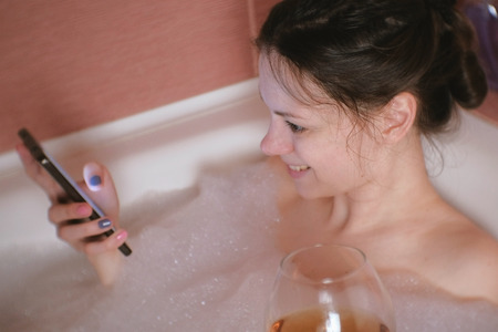 Young beautiful brunette woman takes a bath, drinks whiskey and types a message on mobile phone.