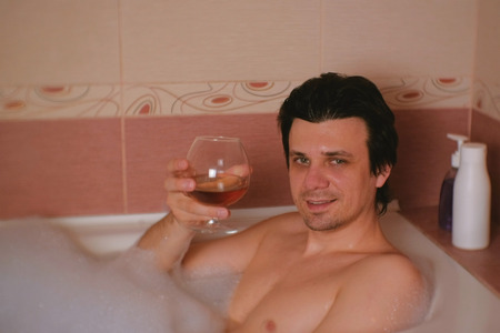 Young man takes a bath with foam and going to drinks whiskey. Smiling and looking at camera.の写真素材