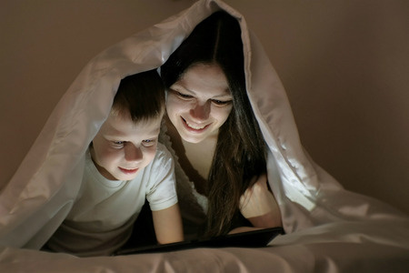 Young woman mom and her son watching a film together on tablet under the blanket.