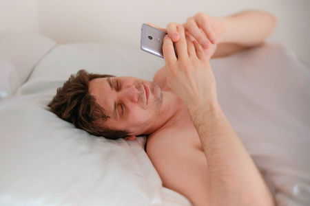 Young brunet handsome man wakes up, and check the time on his mobile phone.の写真素材