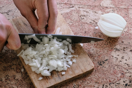 Man gently cuts the onion into small pieces and takes a wooden board with onion from the table. Close-up hands.の写真素材