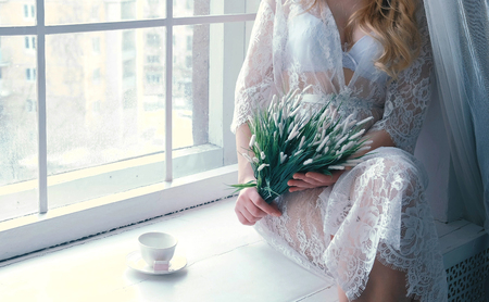 Unrecognizable young slim sexy blond girl in lingerie and boudoir sitting in windowsill in winter. Holds flowers in the hands.の写真素材