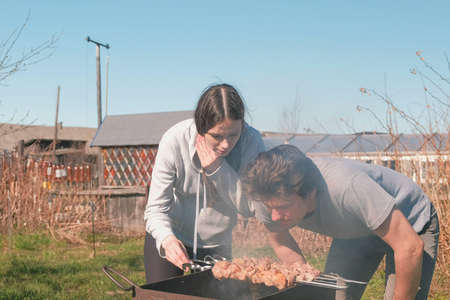 Couple man and woman cook shashlik meat on top of charcoal grill on backyard. Talking and smiling together.の写真素材