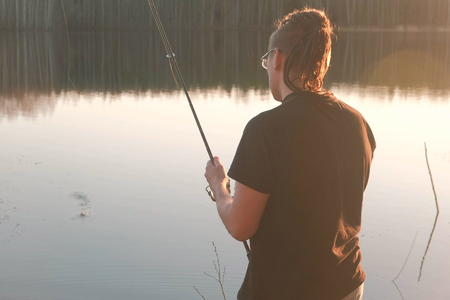 Fisherman on the pond. Young guy with dreads in glasses in a t-shirt fishing fish with rod.の写真素材
