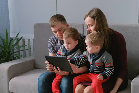 Family with tablet. Mom, dad and two sons twins playing games at tablet sitting in sofa.の写真素材