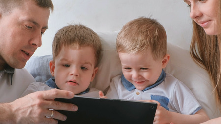 Family with tablet. Mom, dad and two sons twins toddlers looking cartoon at tablet lying on the bed.の写真素材