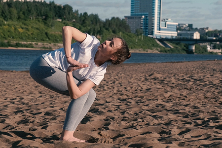 Woman stretching yoga on the beach by the river in the city. Beautiful city view. Utkatasana pose.の写真素材