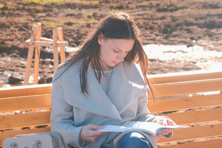 Young beautiful brunette woman student sits on the bench in spring park, reads a book.