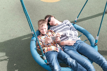 Mom and son swinging on empty webbed swing and playing game on mobile phone together.の写真素材