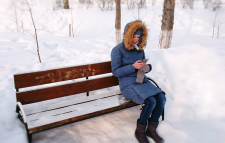 Woman is sitting on bench and browsing mobile phone in winter park in the city in sunny day.の写真素材