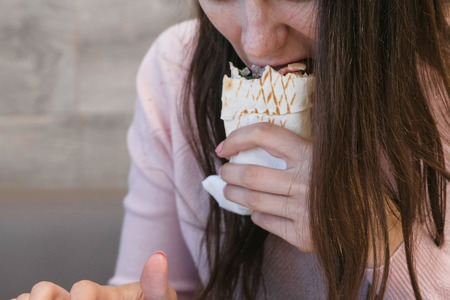 Unrecognizable woman eating danar or Shawarma sitting in a cafe and typing a message at mobile...