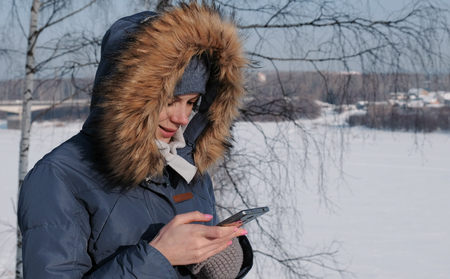 Close-up woman in blue down jacket with fur hood writes messaging in her cellphone in a winter Parkの写真素材