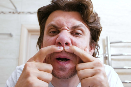 Young man look at mirror and squeezes out a pimple under his nose. Close-up face.