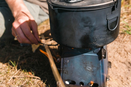 Man inflames metal camp furnace stove on woods outdoors to cook a food. Mens hands close-up.